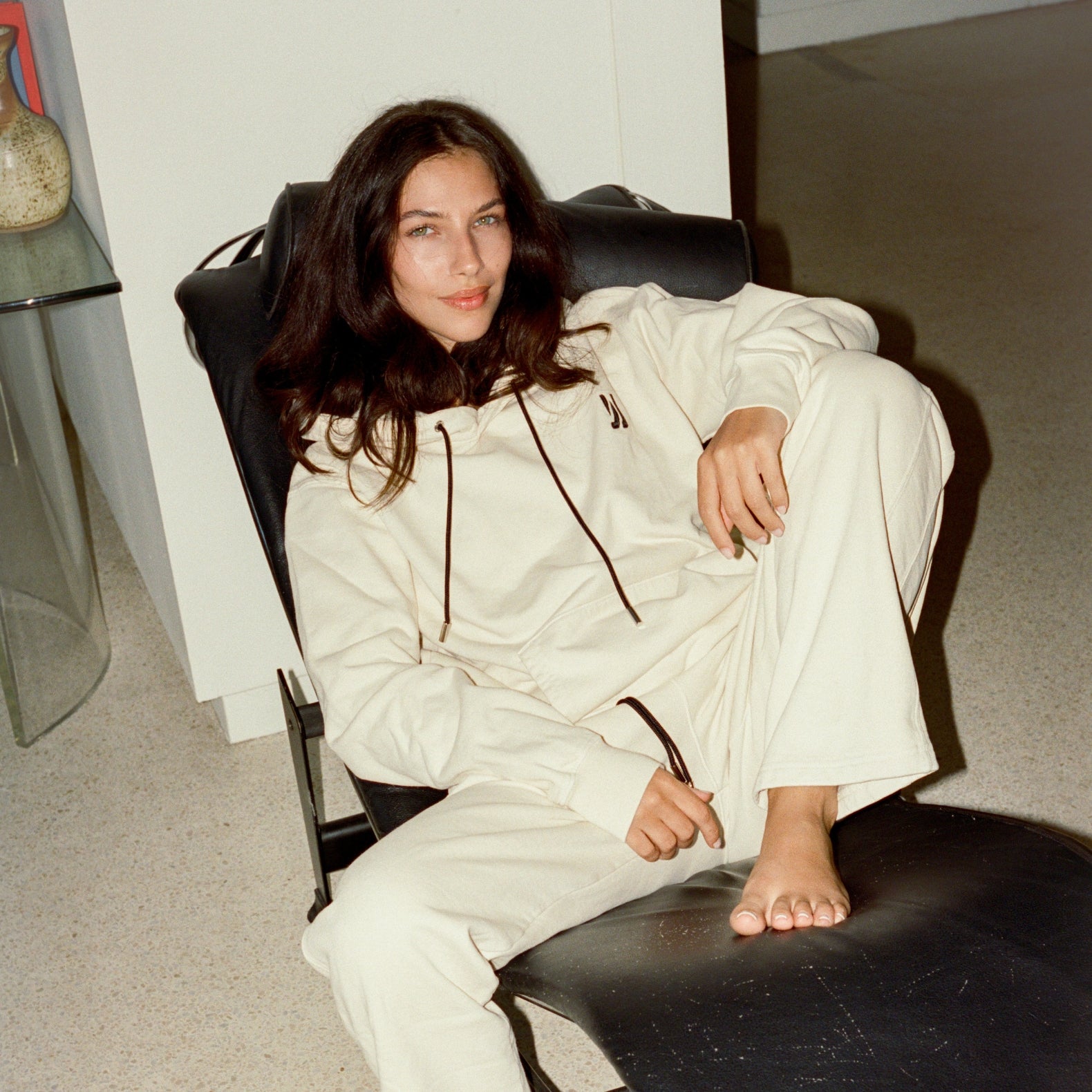 Woman sitting on a black leather sofa wearing Comune Luka Hoodie and Duke sweatpants in both Bone color in a living room. COMUNE LUKA HOODIE | Bone