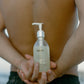 Person holding a bottle of COMUNE Essentials Face Cleanser against his shirtless back