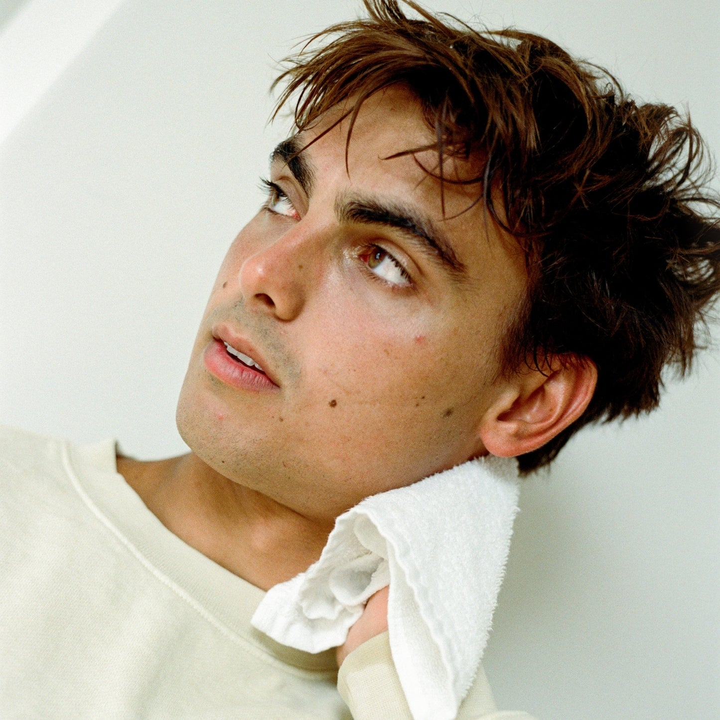 Man with wavy hair wearing a white shirt against a light background, drying his face after using COMUNE Essentials Face Cleanser.