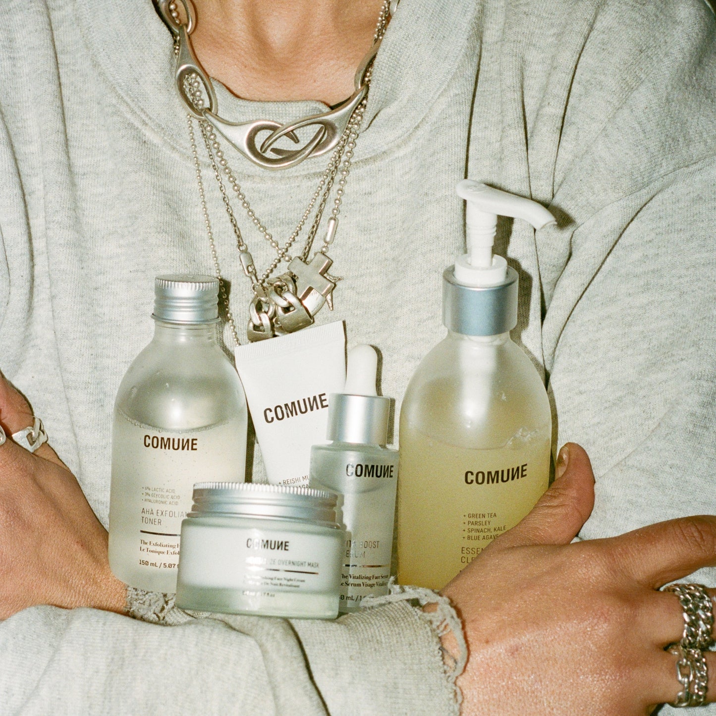 Person holding COMUNE Essentials Skincare products including a gentle face cleanser, moisturizing cream, AHA Exfoliant toner, and other clean skincare products.  Clean, natural, vegan and cruelty-free that are made in Korea.