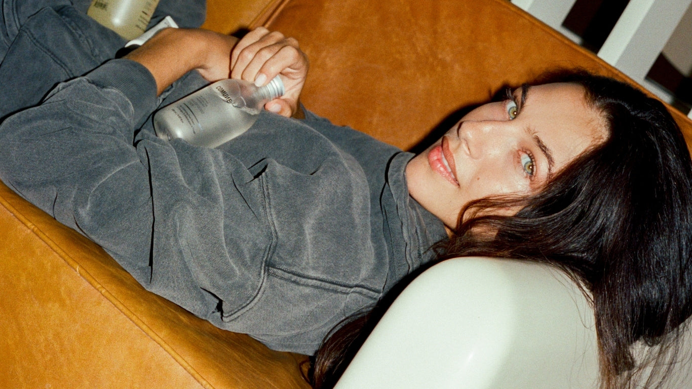 Person lying on a brown leather couch with a neutral expression holding Comune Essentials Skincare Products.  Clean, natural, vegan & cruelty-free.