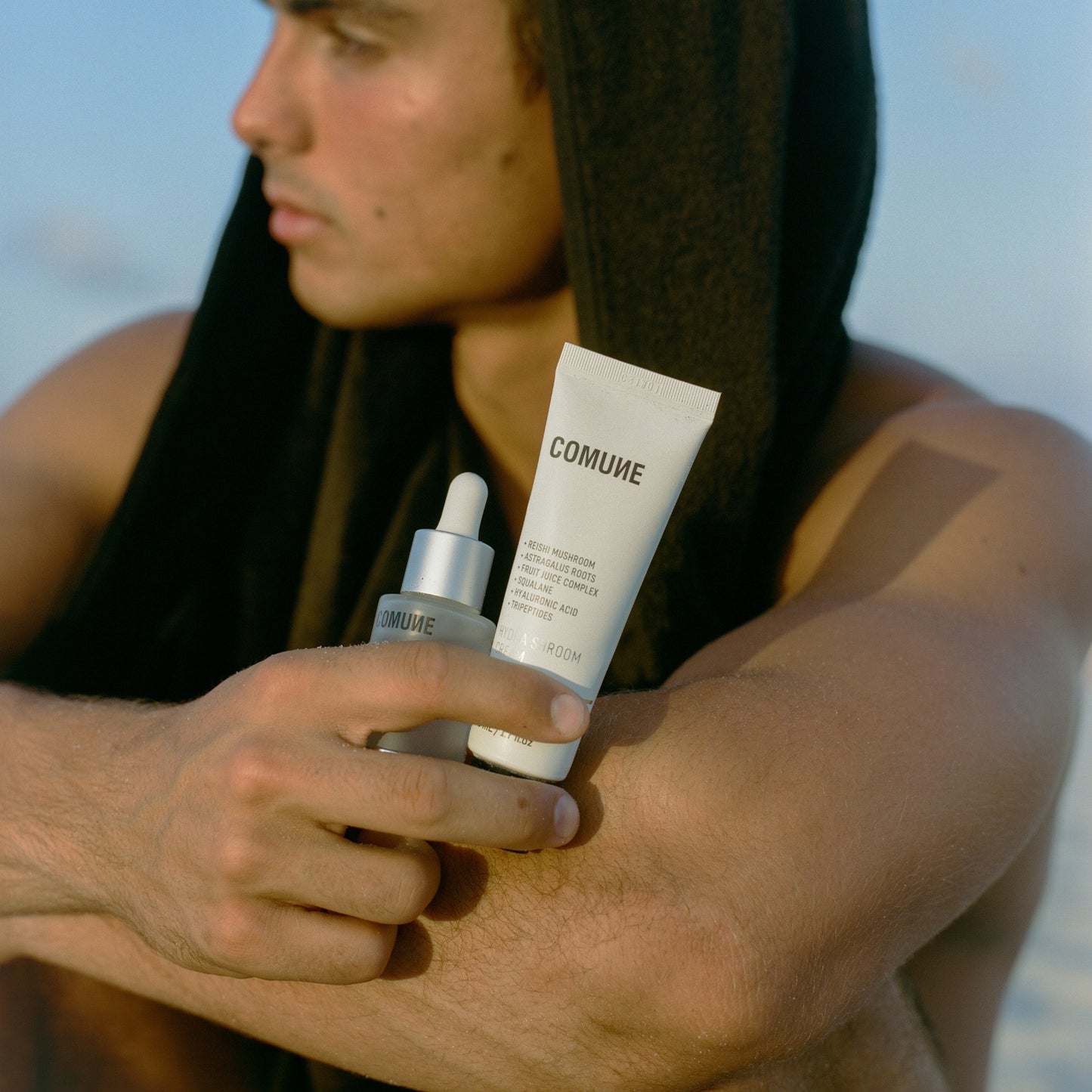 Man holding COMUNE Hydra Shroom Cream products with 'COMUNE' branding against a blurred natural background. Clean, natural, vegan & cruelty-free.