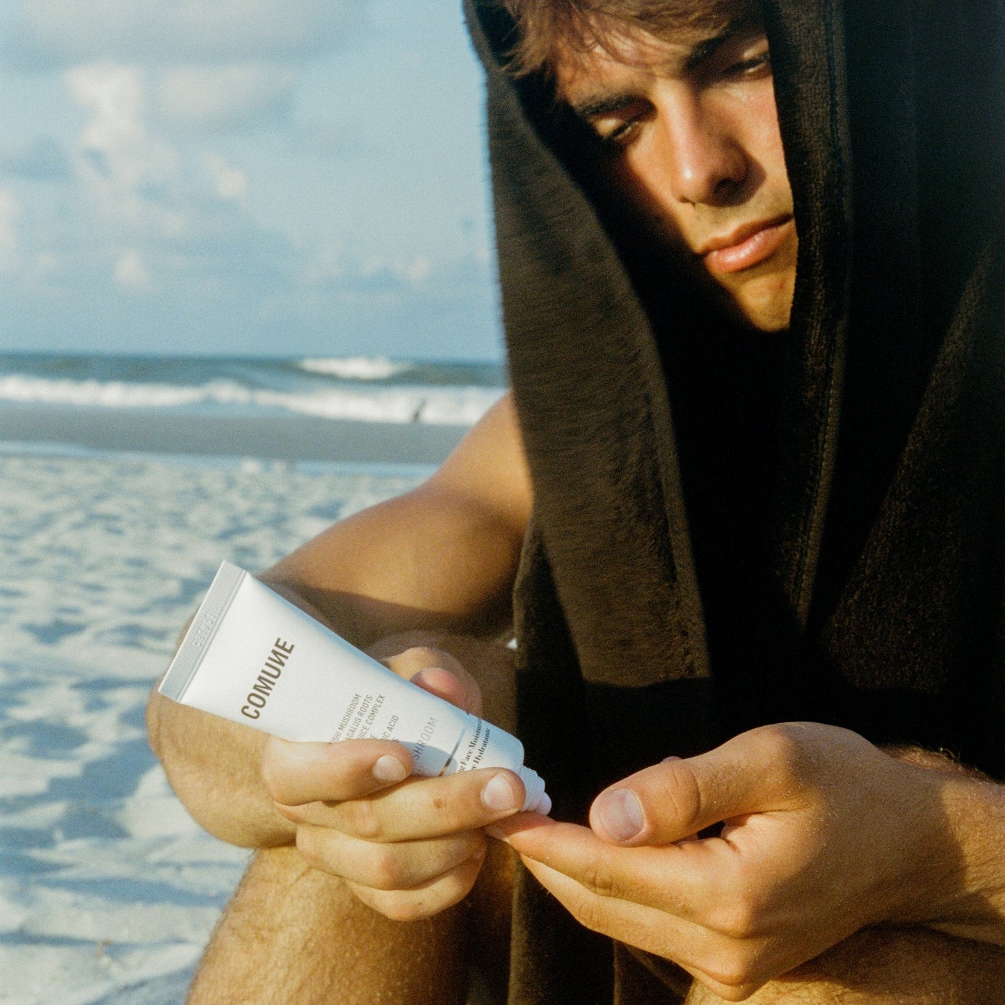 Person on a beach holding a tube of COMUNE Hydra Shroom Cream ready to apply to his face. Clean, natural, vegan & cruelty-free. Made in Korea.