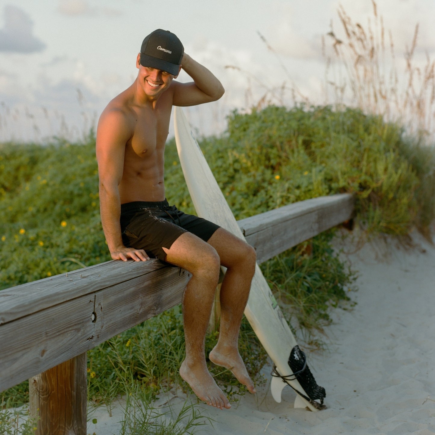 A surfer sitting at the beach wearing COMUNE incursive classic cap headgear and a surf board next to him.