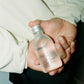 Hand holding a clear bottle labeled 'COMUNE' AHA Exfoliant Toner with a silver cap.