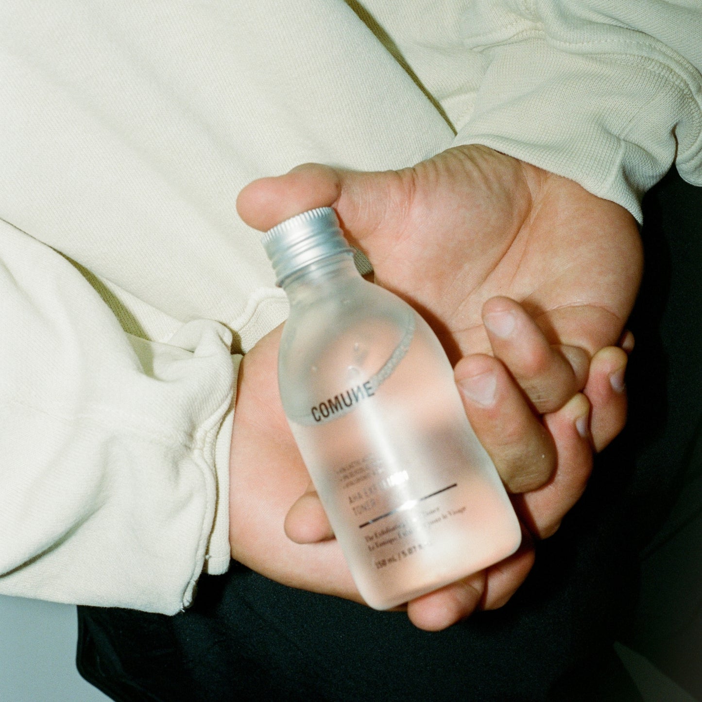 Hand holding a clear bottle labeled 'COMUNE' AHA Exfoliant Toner with a silver cap.