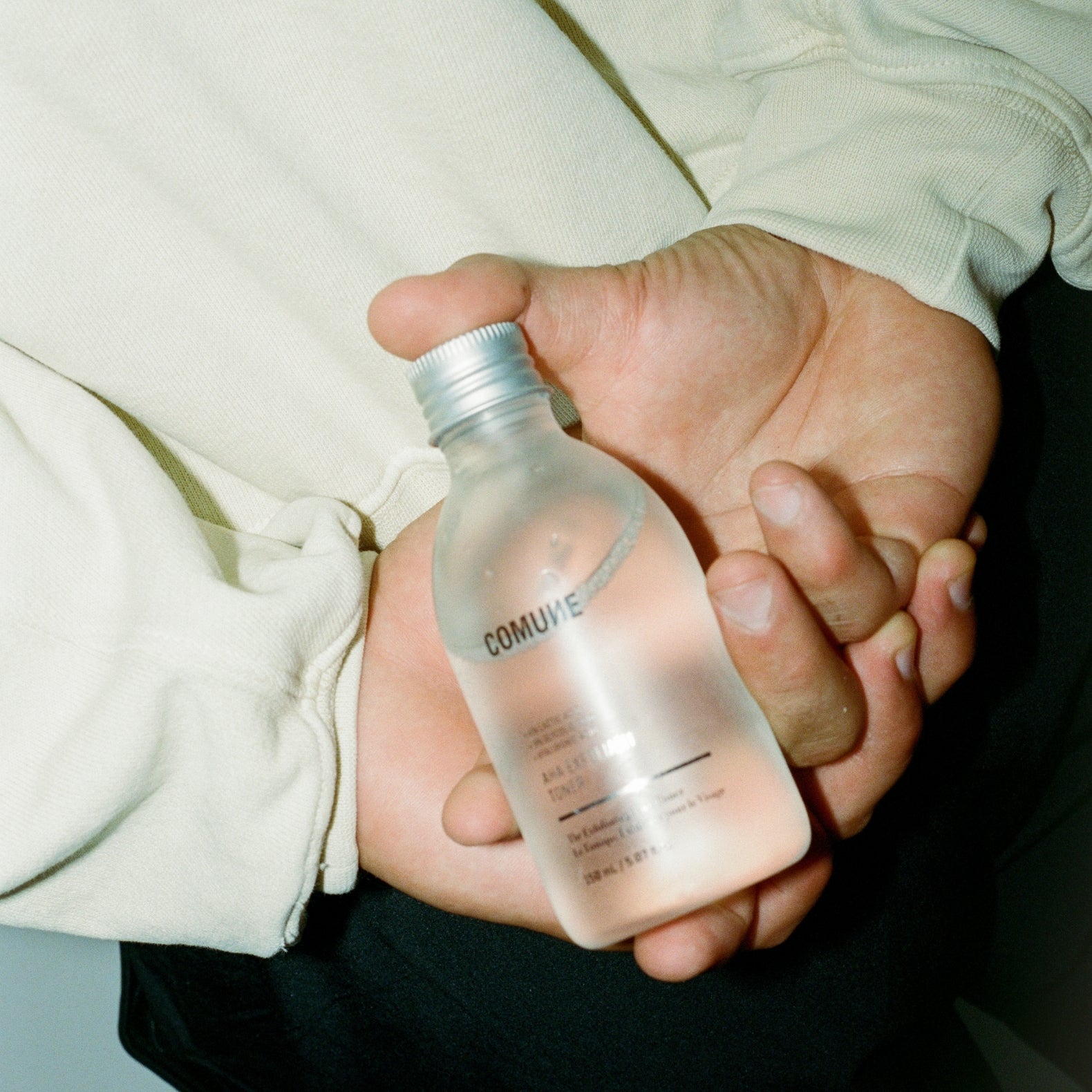 Hand holding a clear bottle labeled 'COMUNE' AHA Exfoliant Toner with a silver cap.
