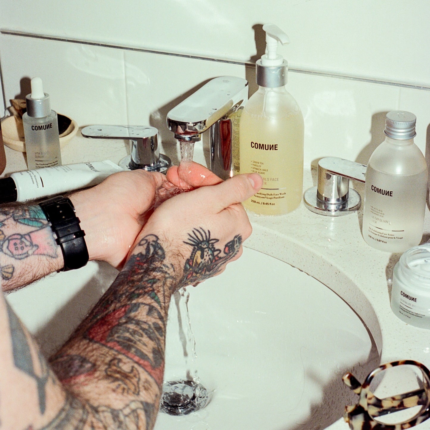 COMUNE artist Ethan washing hands under running water with COMUNE Essentials Skincare products on a sink.