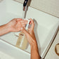 Person holding a bottle labeled 'COMUNE' under running water in a bathroom setting.