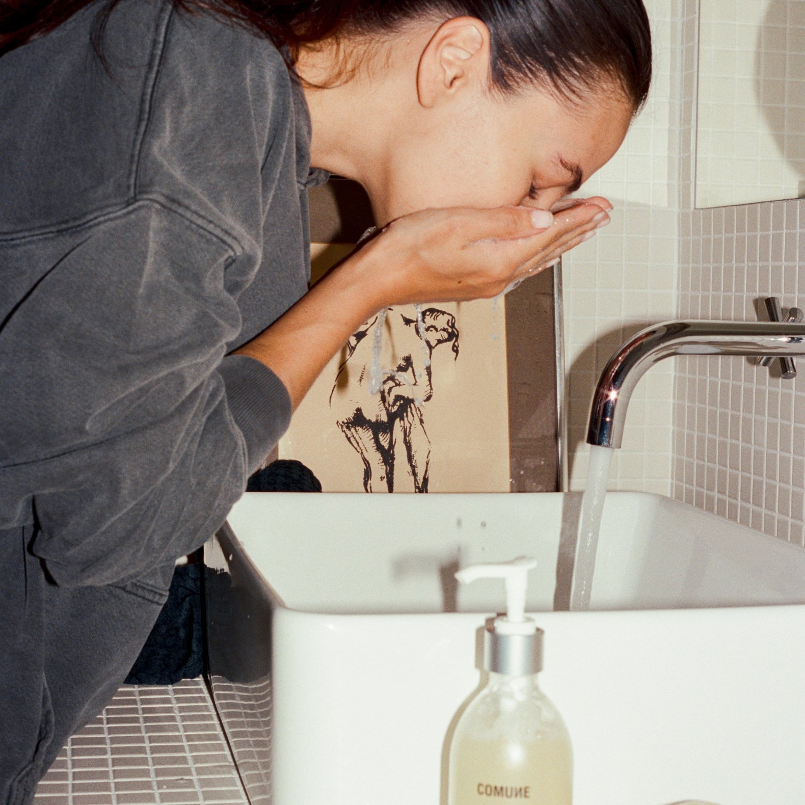 Women washing her face with 'COMUNE' Essentials Face Cleanser in a bathroom sink.