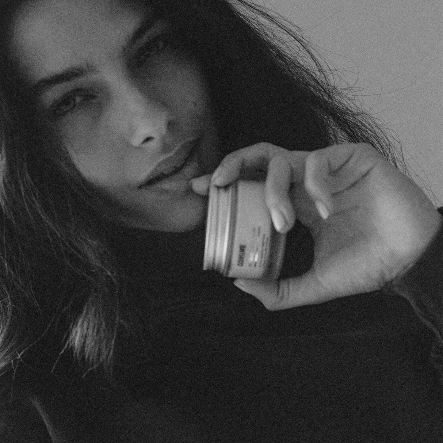 Black and white photo of a person holding 'COMUNE' Revitalize Overnight Mask container, possibly a cosmetic product.