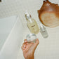 Hand holding a jar of 'COMUNE' skincare product with bottles in the background on a tiled bathroom floor.