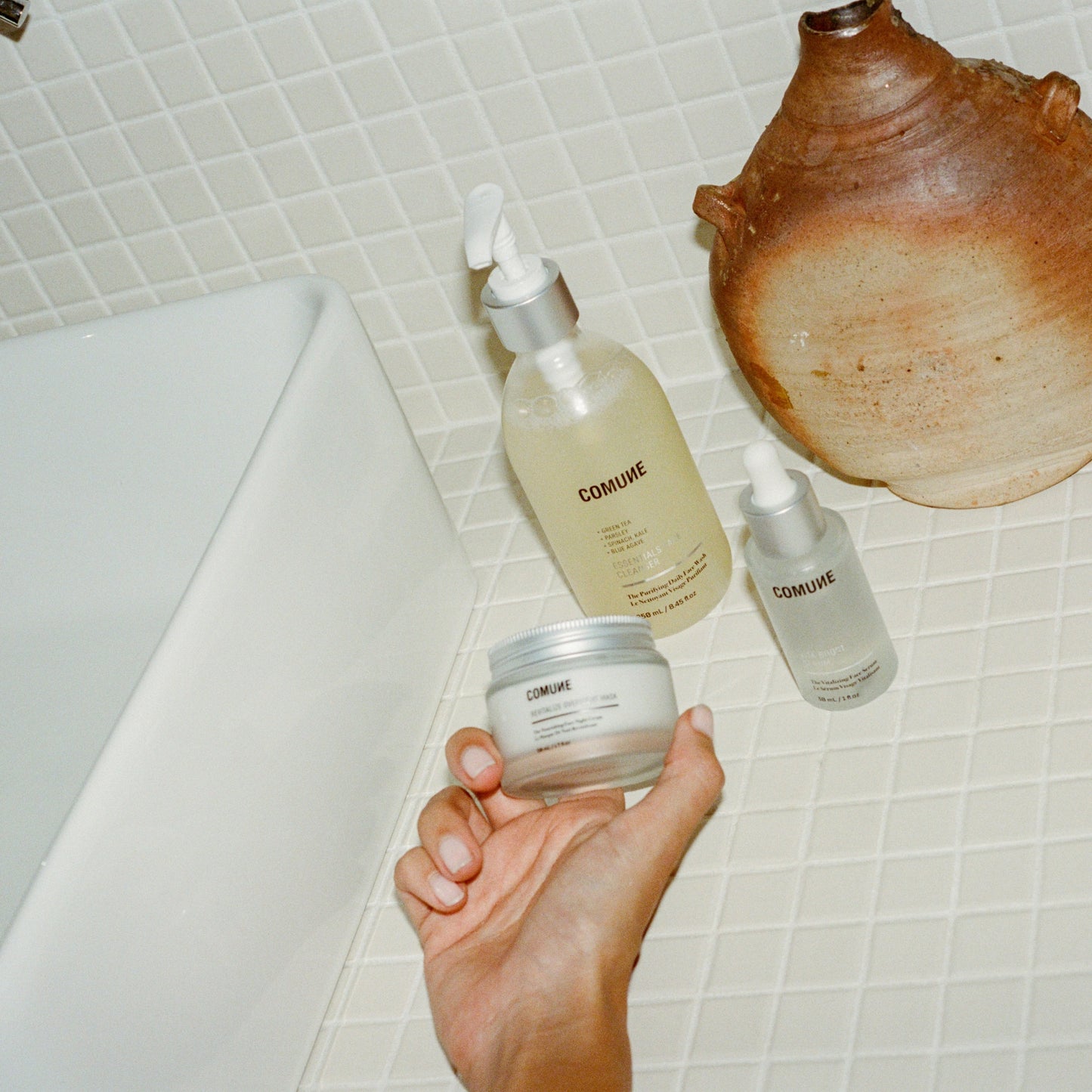 Hand holding a jar of 'COMUNE' skincare product with bottles in the background on a tiled bathroom floor.