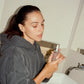 Woman in a bathroom holding a "COMUNE' vita boost serum skincare product