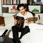 COMUNE artist Brandon playing guitar on a couch with bookshelves in the background
