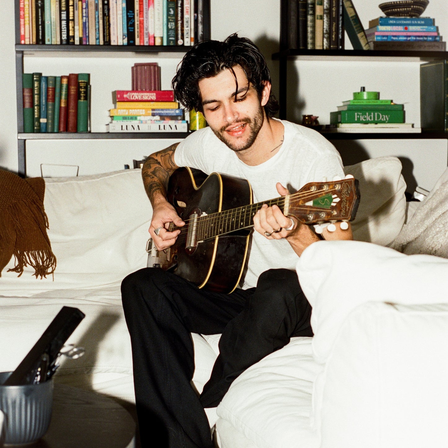 COMUNE artist Brandon playing guitar on a couch with bookshelves in the background