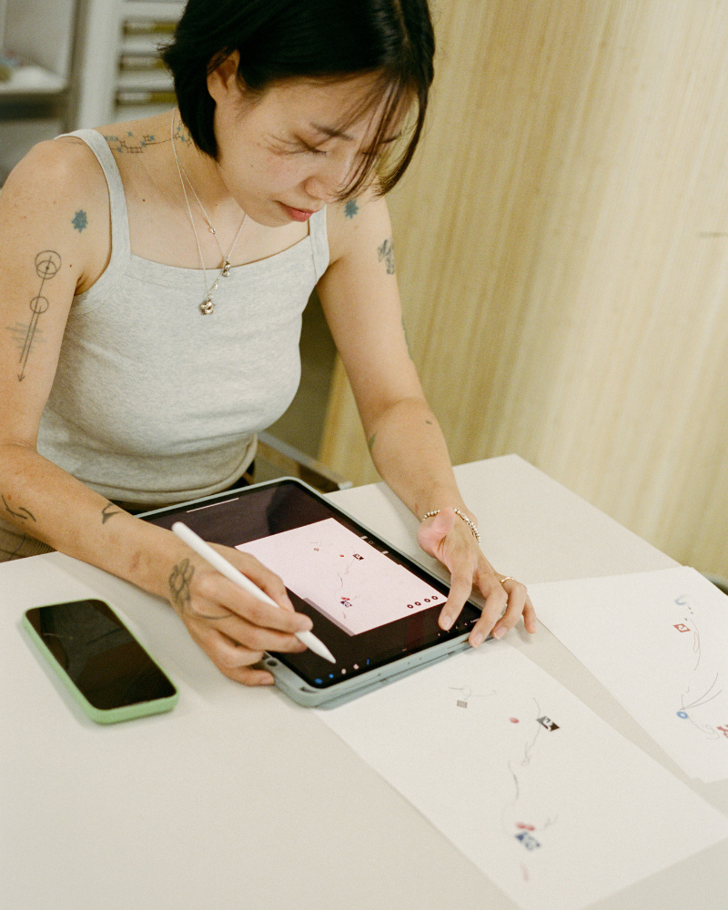 COMUNE artist Odni using a stylus on a tablet with a phone beside them on a table as she work on creating tattoo drawing