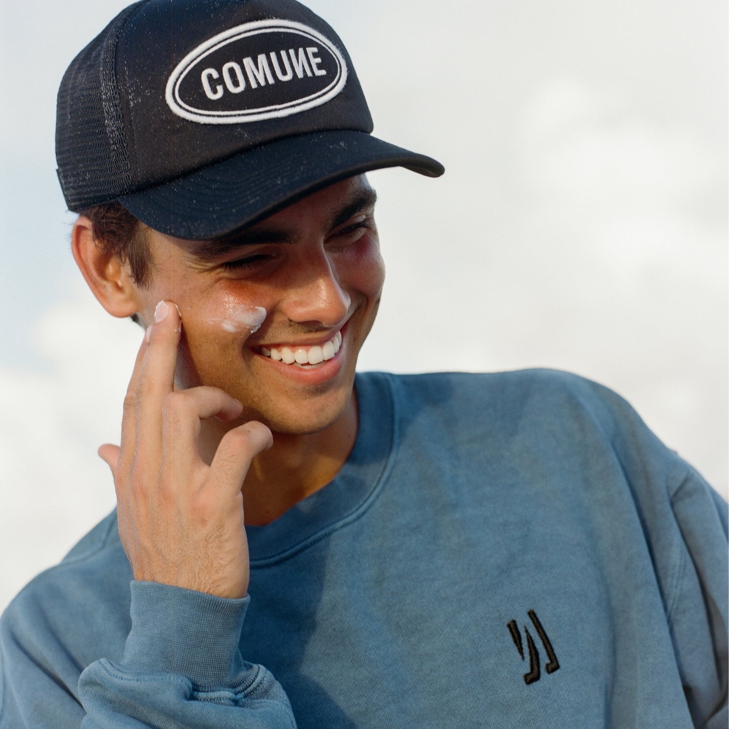 Man wearing a black cap with 'COMUNE' logo and marine COMUNE Duke Crew sweatshirt, applying sunscreen.