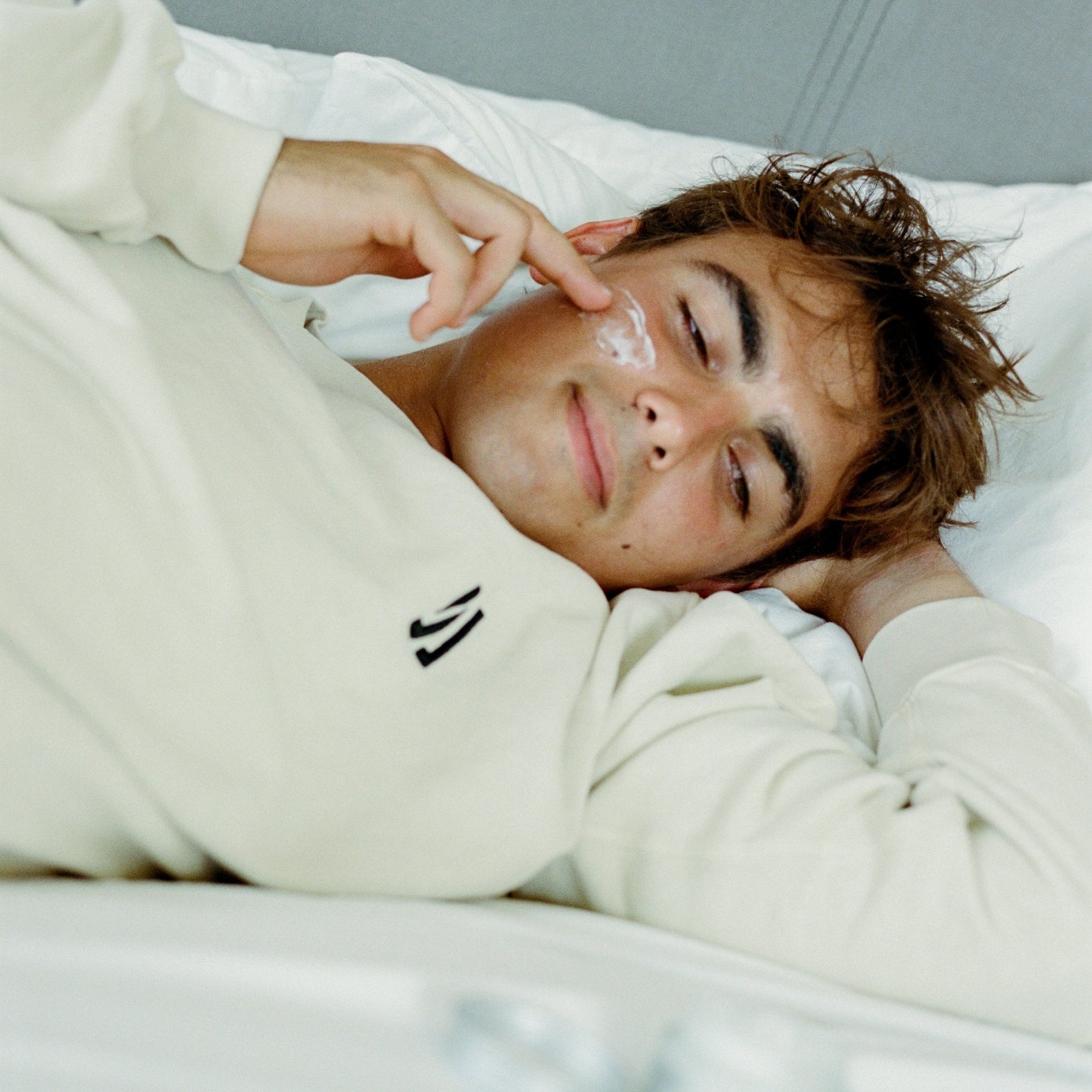Person lying in bed wearing a COMUNE Duke Crew sweatshirt with a logo, looking relaxed applying comune hydra shroom cream.