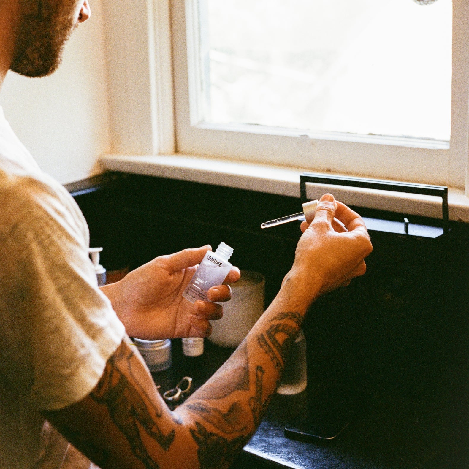 COMUNE artist Brandon holding a comune's vita boost serum and a dropper near a window.
