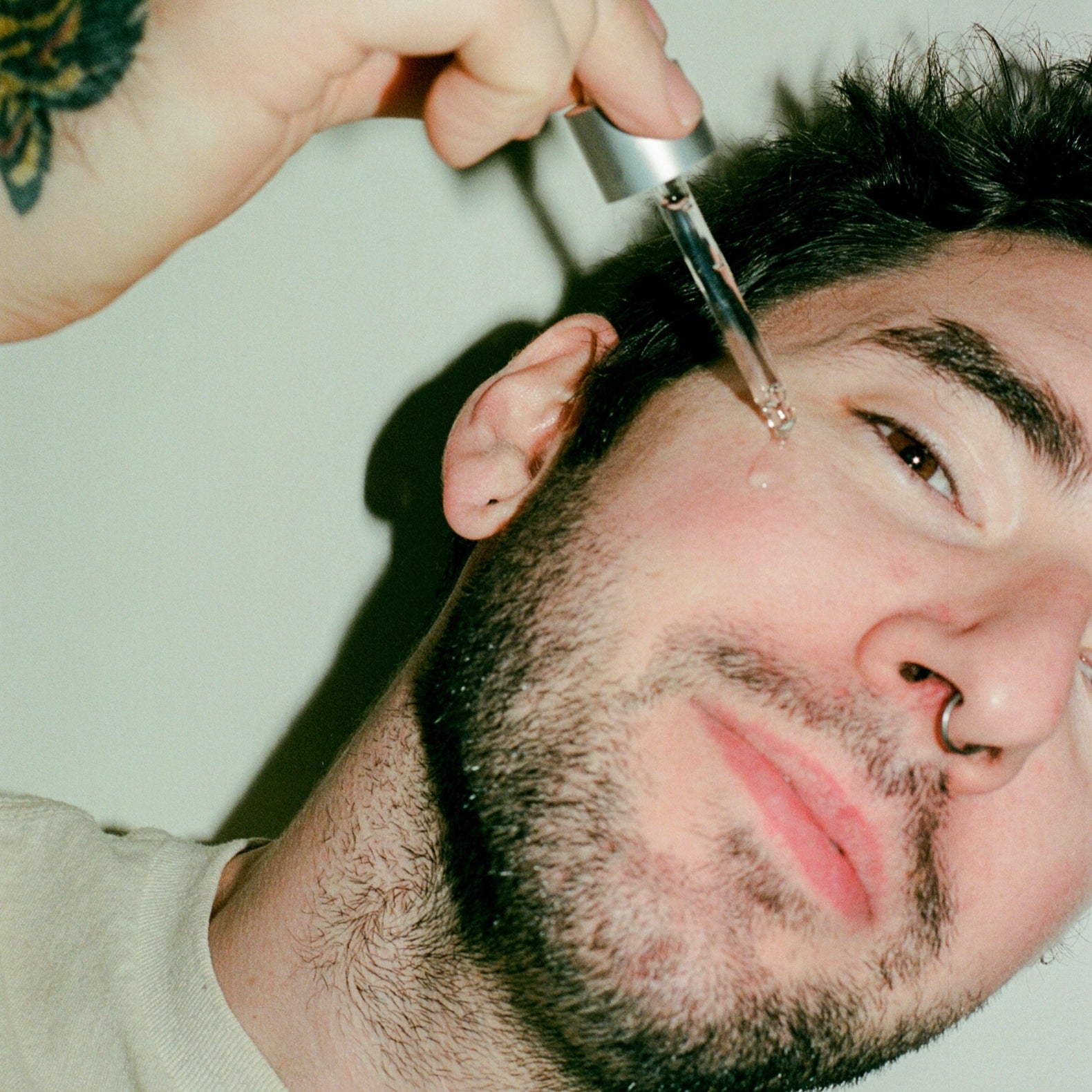 Person applying a comune vita boost serum to their face against a neutral background.  This clean and natural serum is made from powerhouse vegan ingredients that is packed of antioxidants, vitamins, peptides, and hydrating ingredients for anti-aging and nourishing properties.