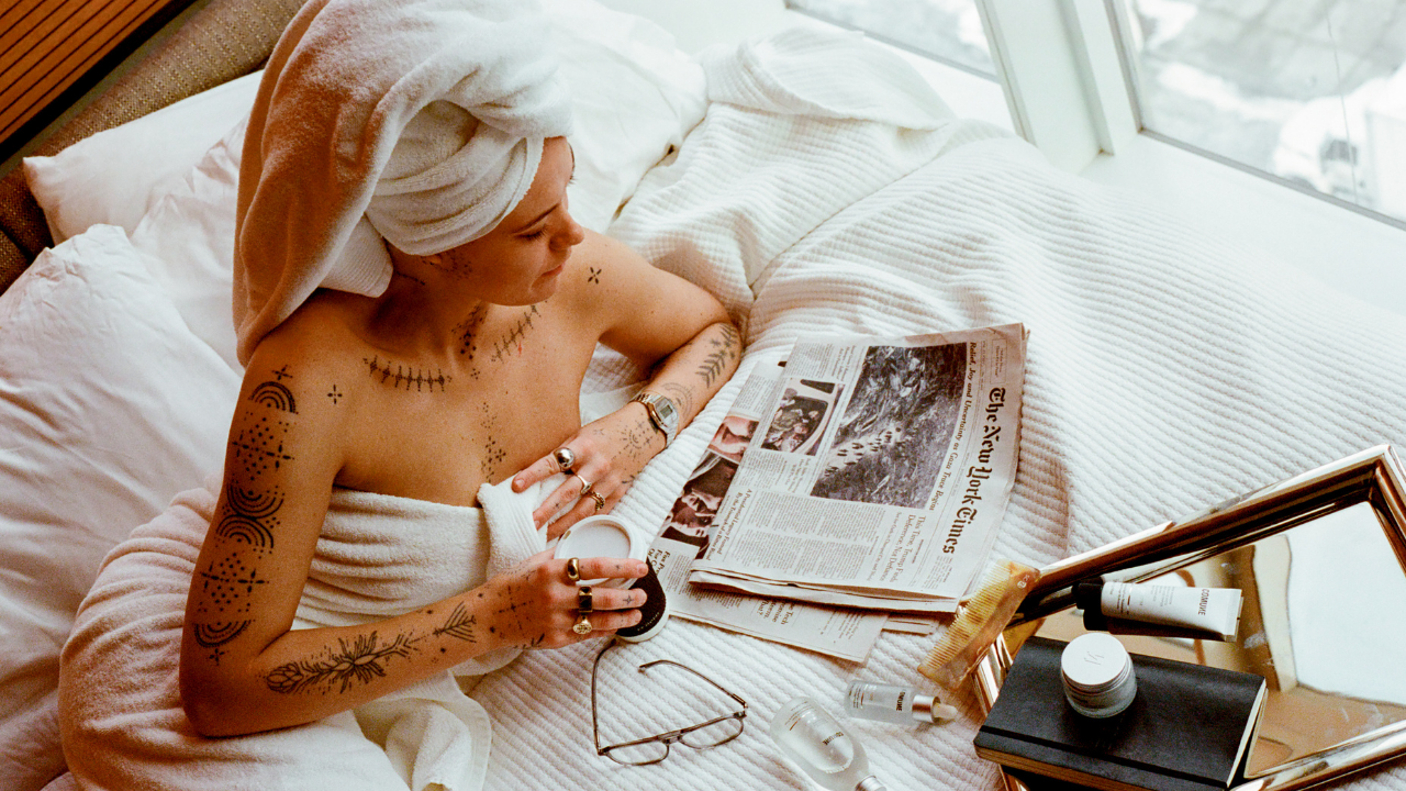 COMUNE model with a towel on her head reading a newspaper in bed with 'COMUNE' Essentials Skincare products laying on the bed.